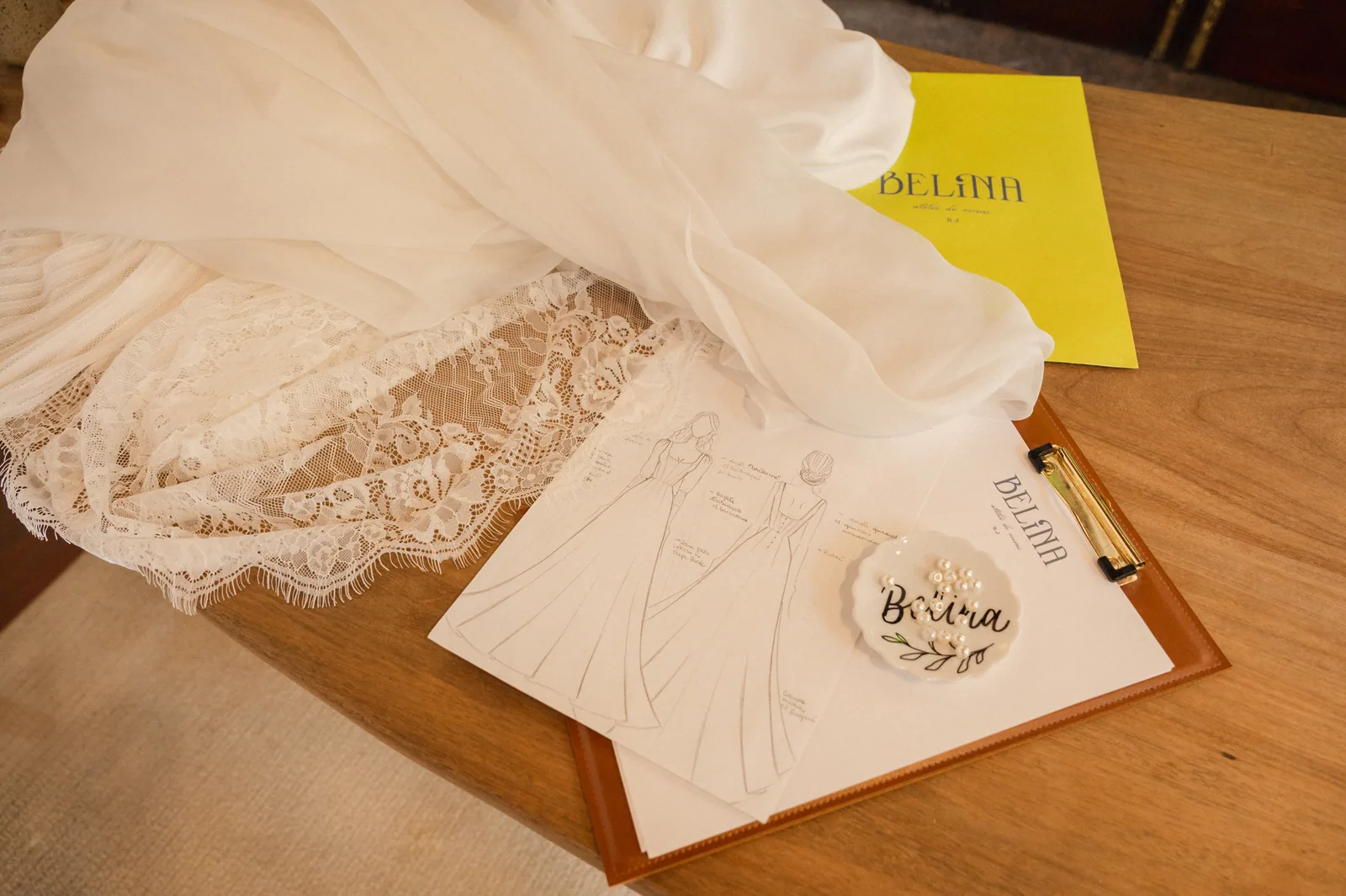 Croqui de vestido de noiva sobre a prancheta da Belina, ao lado de renda e seda na mesa do ateliê