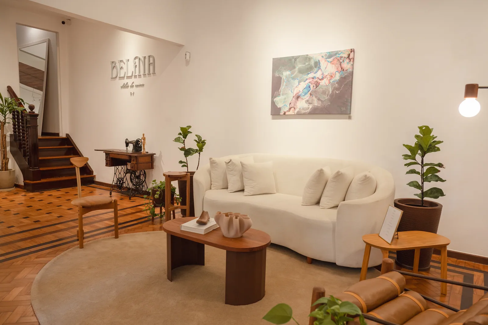 Sala da Casa Belina com sofá curvo claro, poltrona de couro, escada de madeira, máquina de costura antiga e o letreiro do ateliê na parede